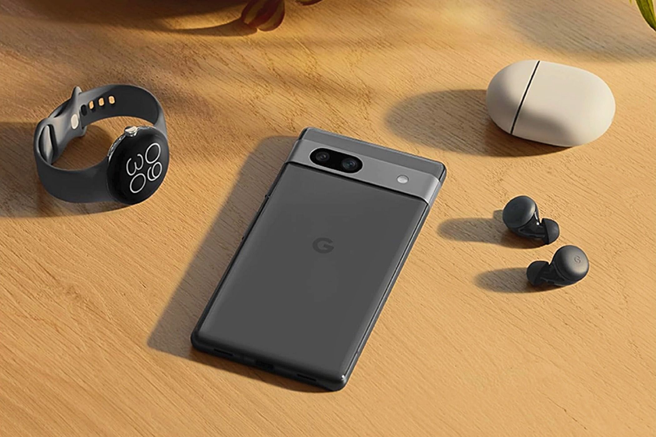 A Google Pixel 7a on a desk next to a smartwatch, earbuds, and charging case.