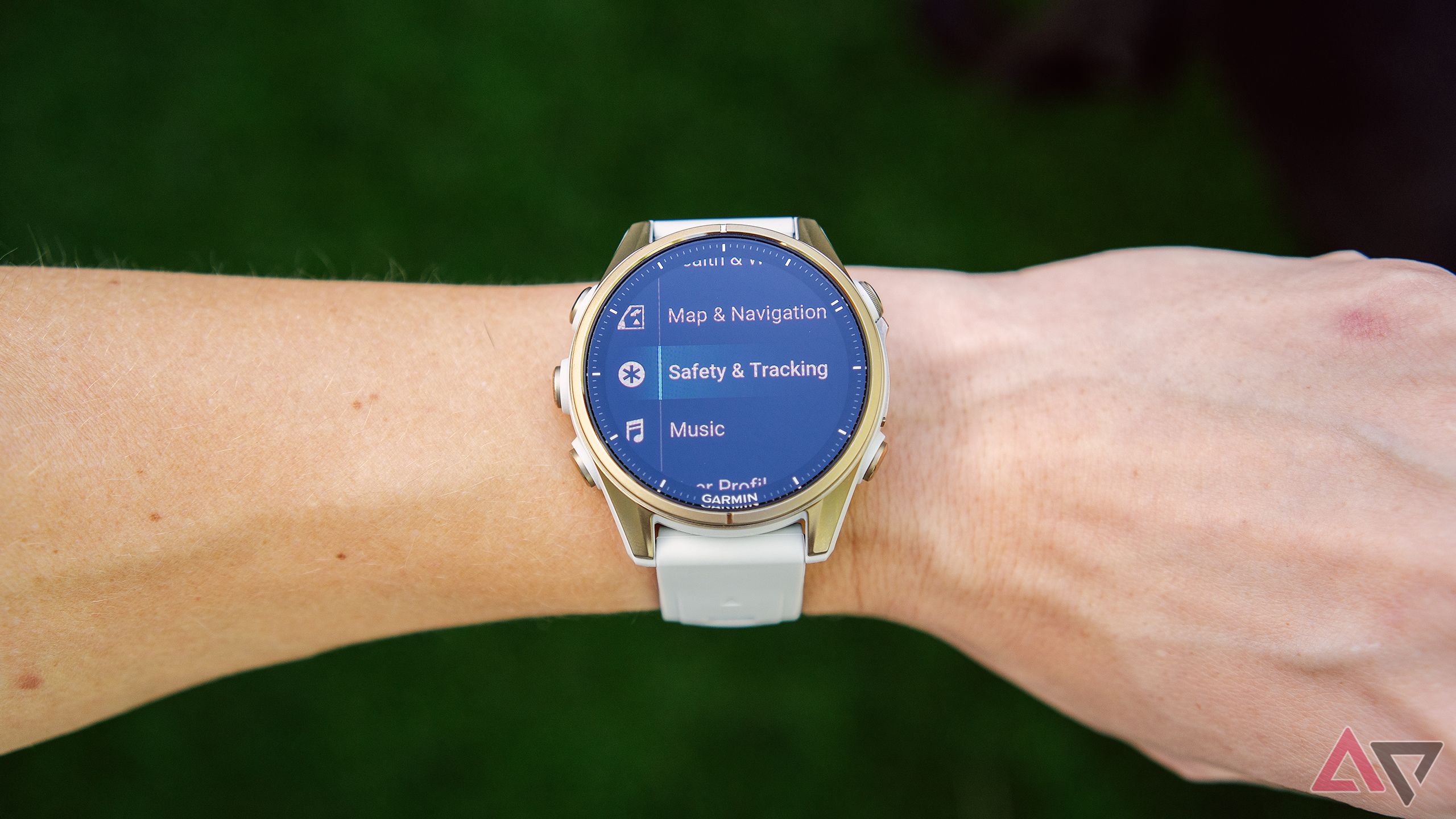 A wrist with a Garmin Fenix 8 AMOLED and the Safety & Tracking features on the display is held above a blurred green background. 